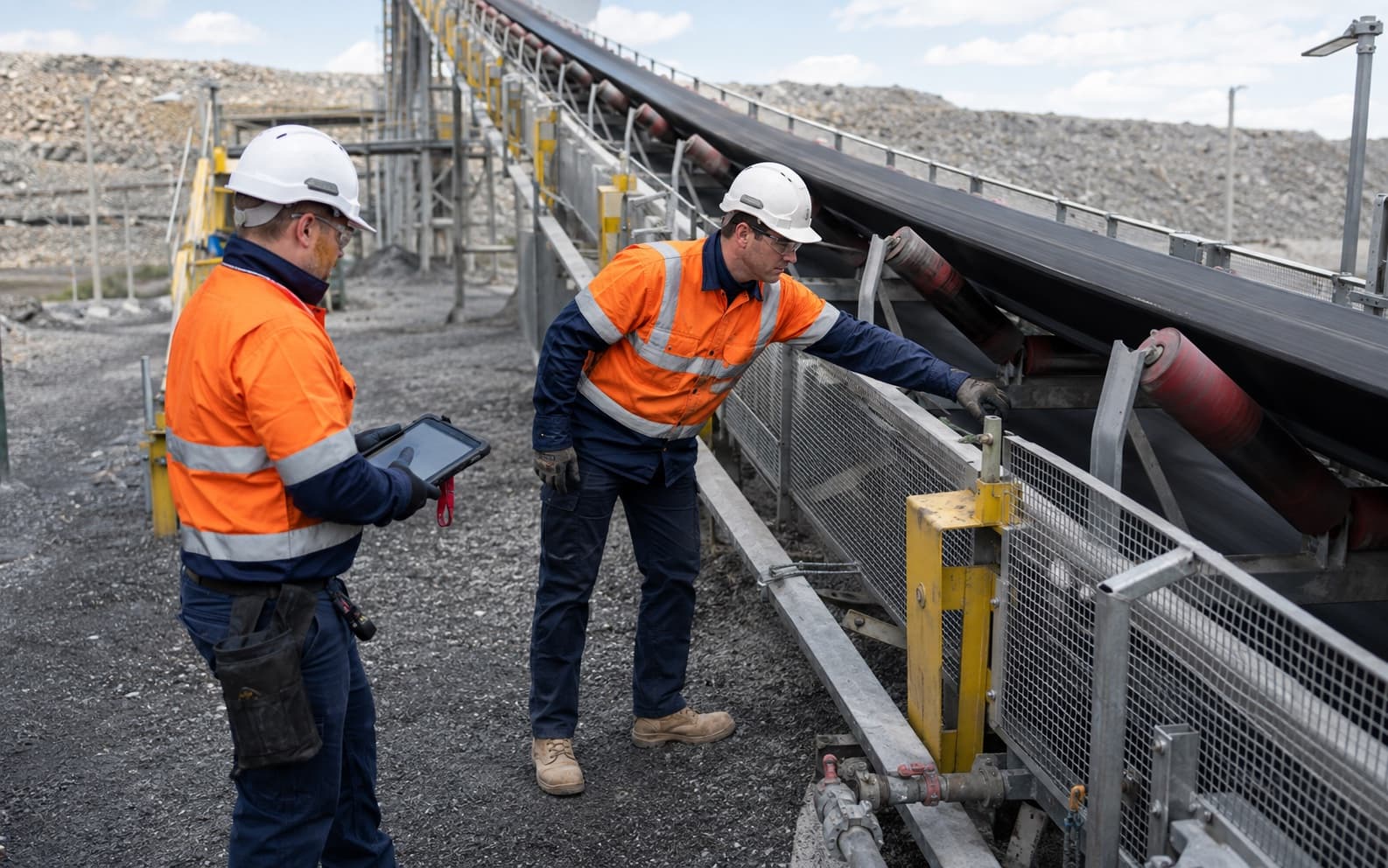 Two conveyor maintenance technicians inspect belt tracking and idler condition on an isolated quarry conveyor